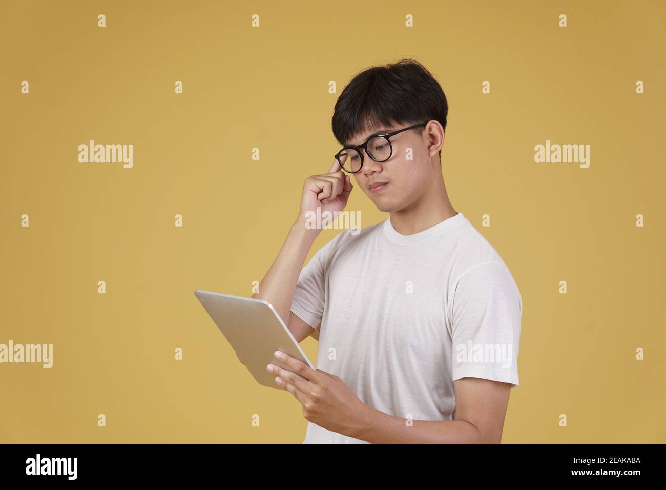 un jeune homme asiatique attentionné s'habille en pensant à la manière décontractée en utilisant une tablette numérique isolée sur fond jaune de studio. Banque D'Images