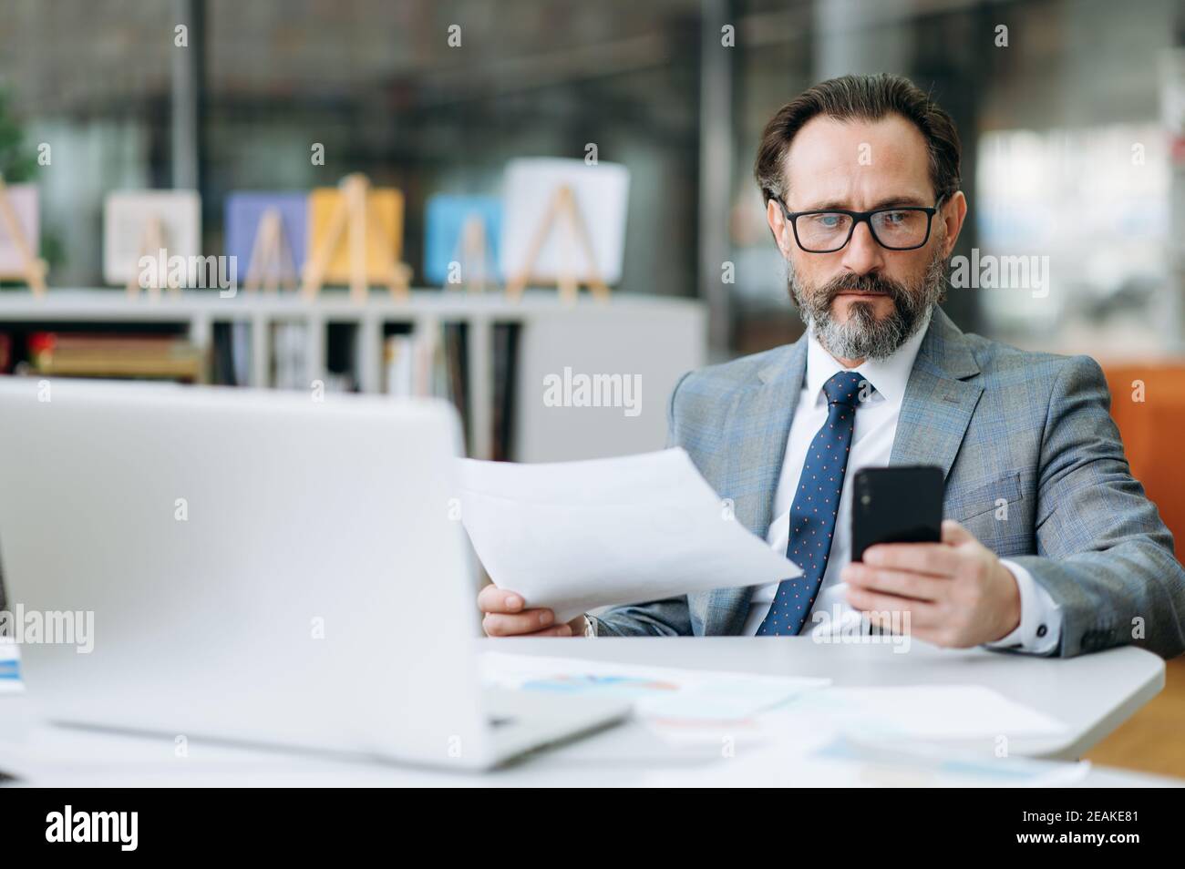 Un homme sérieux d'âge moyen est assis à la table dans un bureau moderne, travaille avec des documents. Un homme d'affaires concentré utilise un smartphone, navigue sur Internet, recherche des idées de projet ou de démarrage Banque D'Images