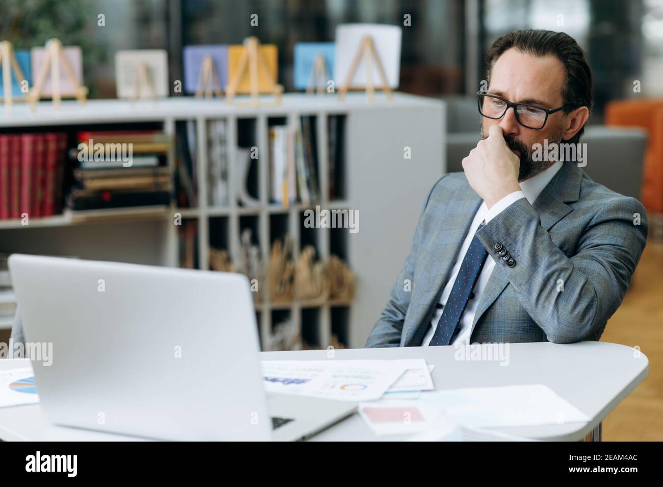 Homme d'affaires d'âge moyen vêtu d'un costume élégant, concentré sur un projet sérieux, assis au bureau dans un bureau moderne. Un responsable masculin spécialisé utilise un ordinateur portable, réfléchit à la stratégie de l'entreprise Banque D'Images