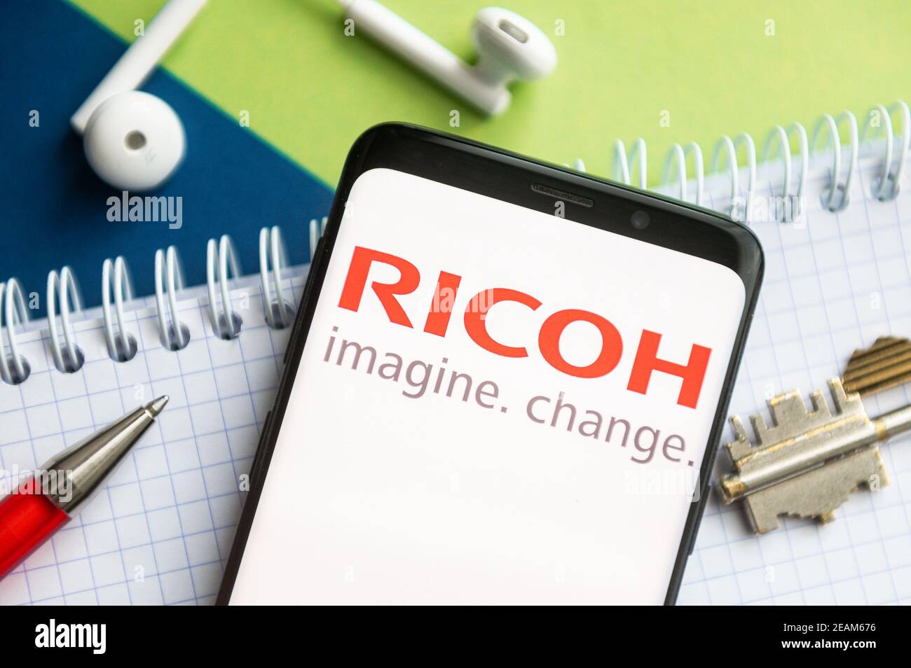 Dans cette illustration, un logo Ricoh apparaît sur un smartphone avec un stylet, une touche, un livre et des casques en arrière-plan. Banque D'Images