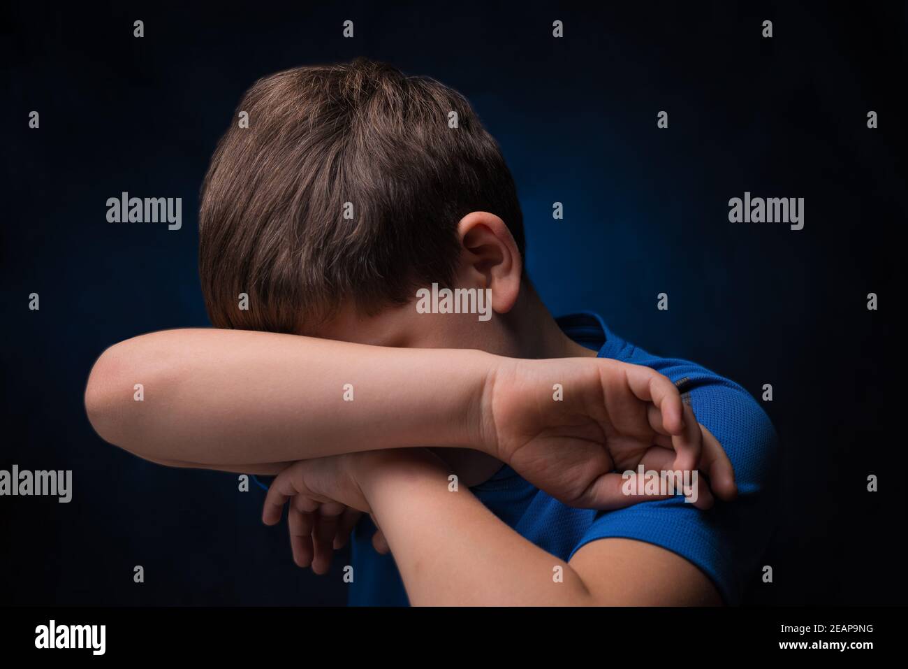 pleurer un adolescent européen avec des cheveux brun clair, portant un t-shirt de sport bleu, couvrant son visage avec la main sur un arrière-plan isolé Banque D'Images