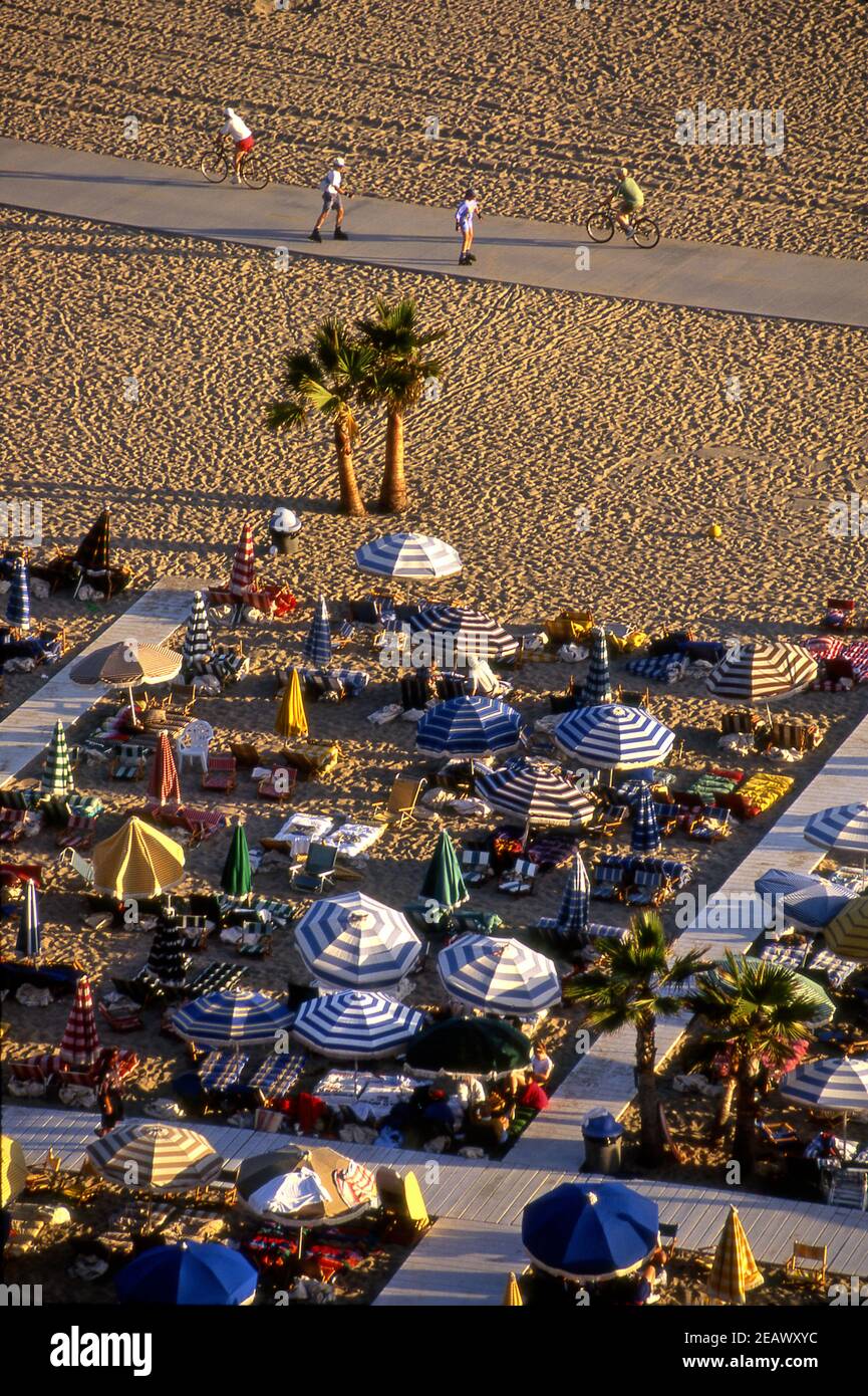 Des vélos et des patineurs sur la piste cyclable de la plage en passant devant un club de plage avec des parasols colorés à Santa Monica, en Californie Banque D'Images