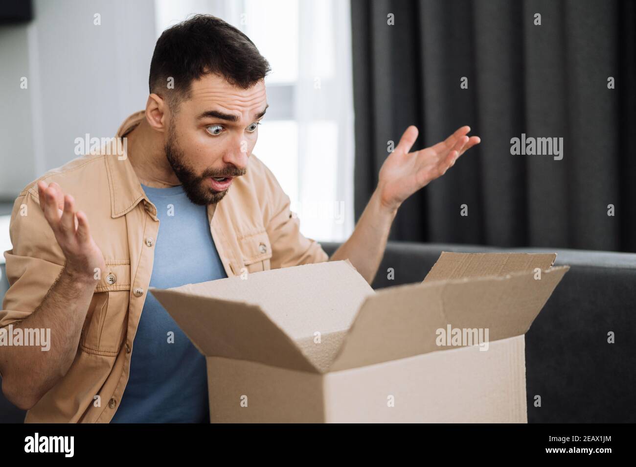 Livraison à domicile. Un jeune homme choqué s'assoit sur le canapé,  déballant son colis. Surprise caucasien mâle a reçu une mauvaise commande  ou une boîte de carton vide. Mécontent g Photo Stock -