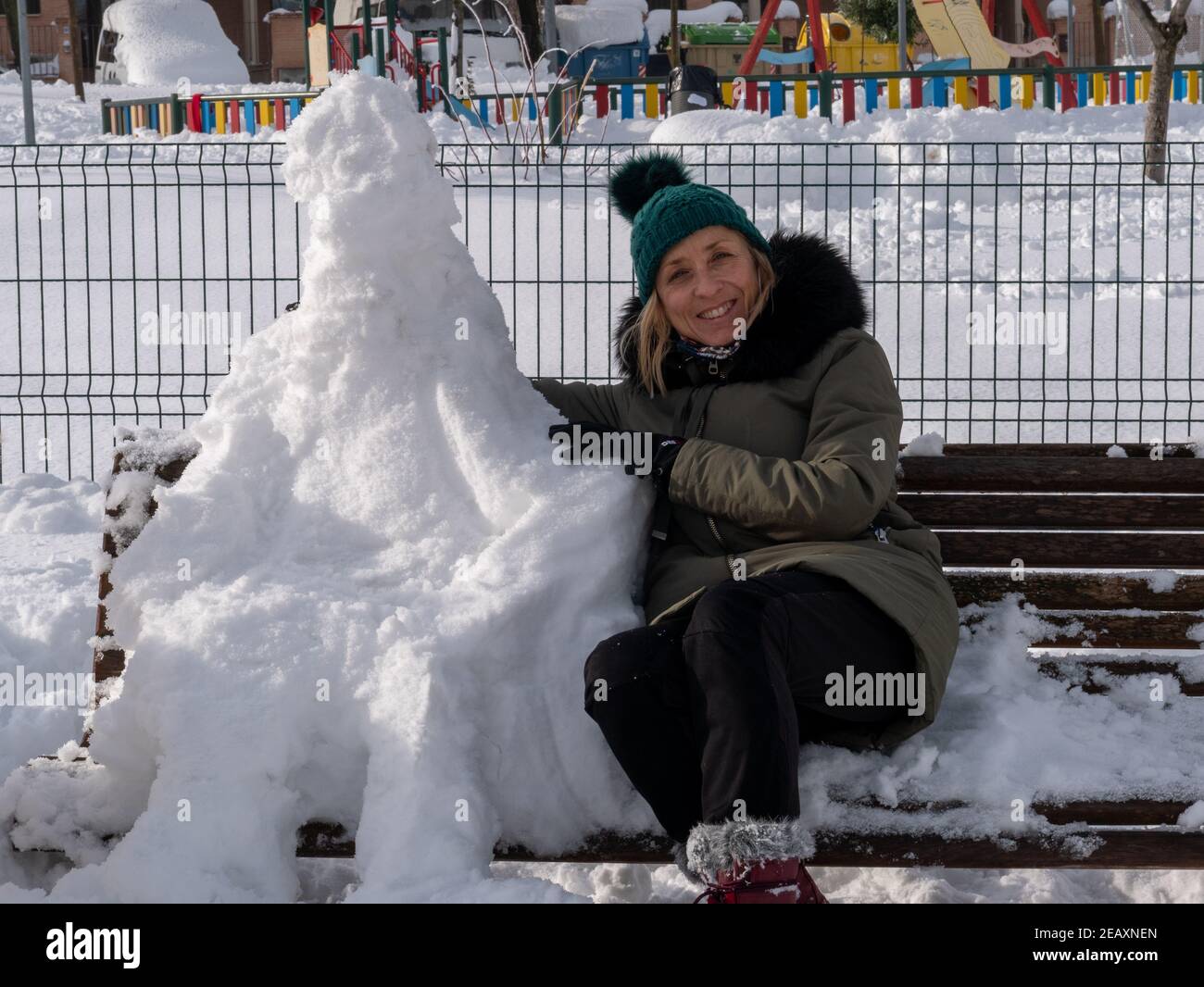 Blonde gaie d'âge moyen dans un manteau assis sur un banc à côté d'un bonhomme de neige dans un parc Banque D'Images