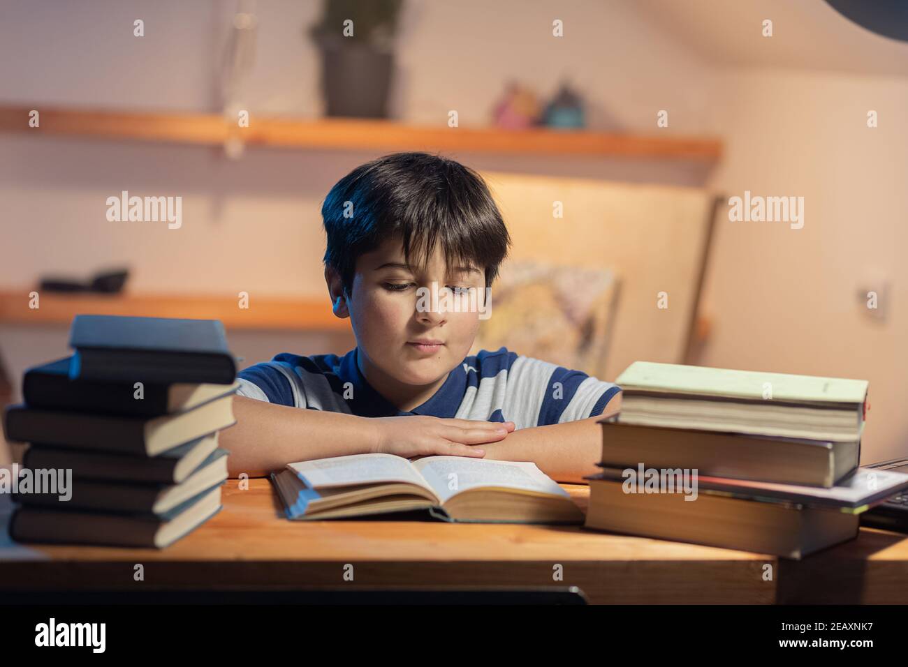 Portrait d'un garçon qui lit. Il est assis dans sa chambre meublée de piles de livres et il est attentif à la lecture. Banque D'Images