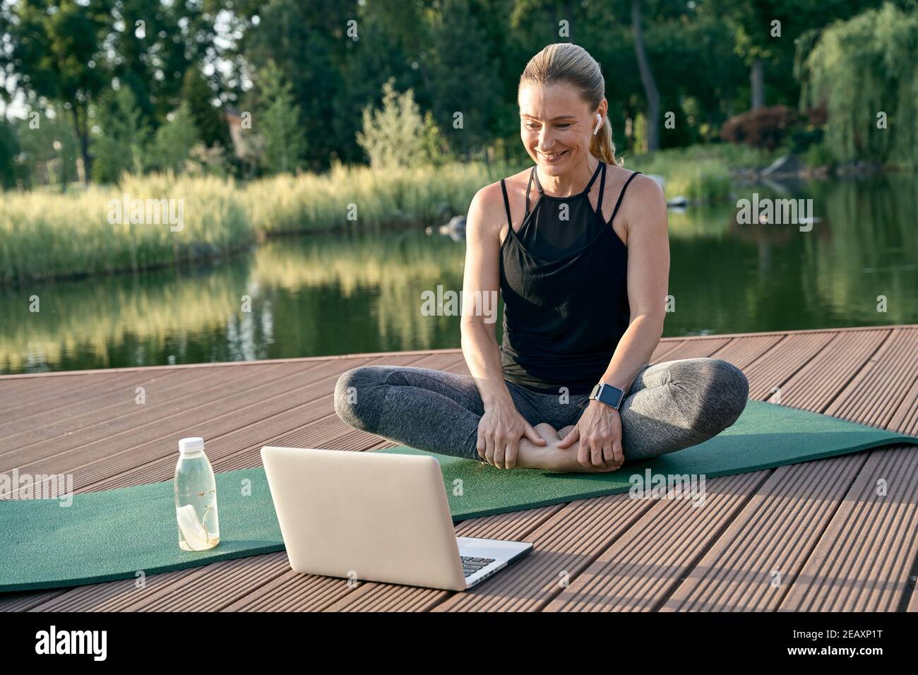 Femme sportive et belle qui a l'air heureuse, regardant un tutoriel sur un ordinateur portable tout en faisant du yoga sur un tapis dans la nature pendant une journée ensoleillée à l'extérieur Banque D'Images