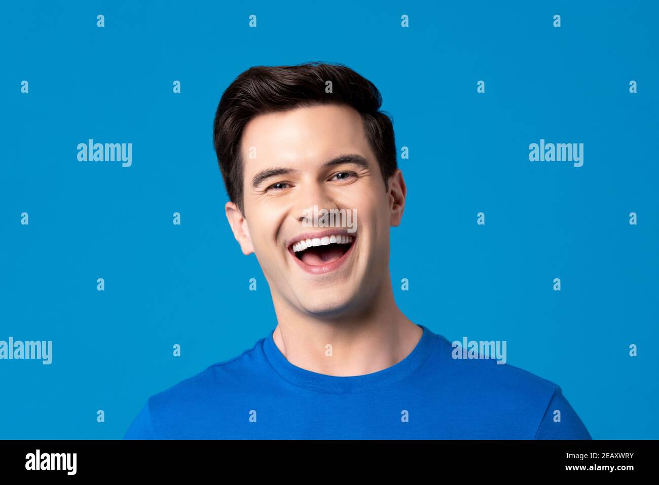 Beau et heureux jeune homme caucasien en t-shirt bleu clair rire isolé sur fond de studio bleu clair frais Banque D'Images