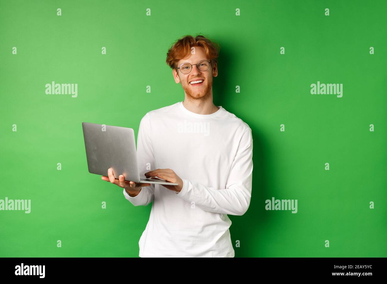 Joyeux freelance dans des verres souriant à l'appareil photo, travaillant sur un ordinateur portable tout en se tenant debout sur un fond vert Banque D'Images