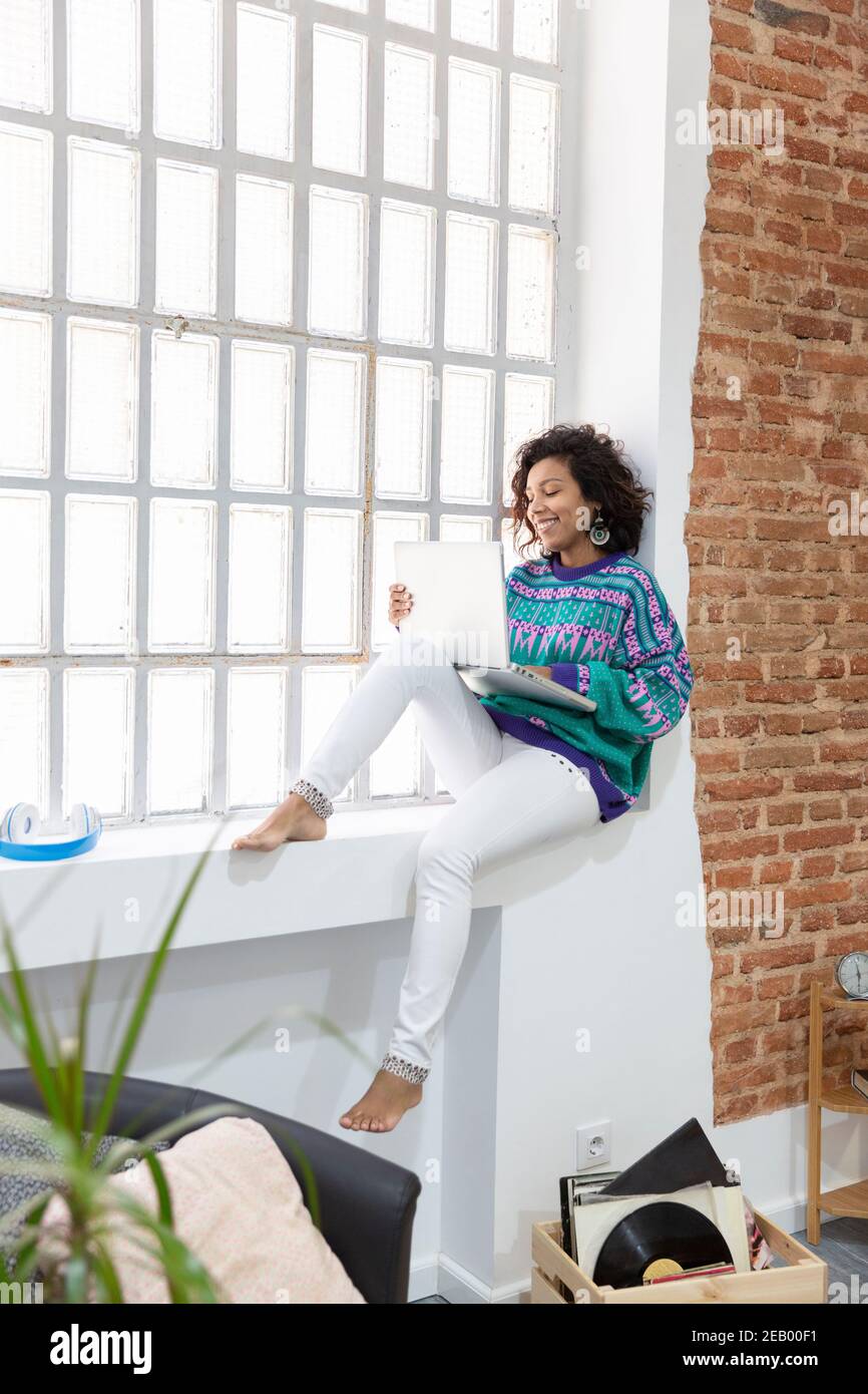 Jeune femme vêtue de façon décontractée travaillant sur un ordinateur portable tout en étant assise sur le seuil de la fenêtre à la maison. Concept « travail à domicile ». Banque D'Images