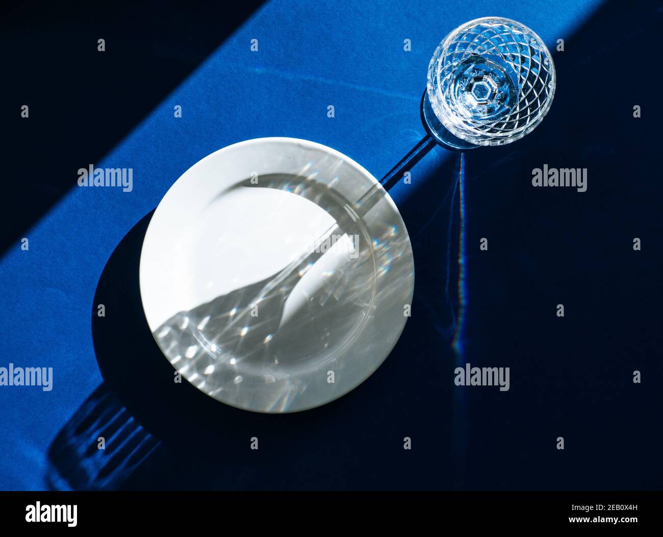 Table avec plaque blanche vide et verre cristal isolé sur fond bleu foncé, ombres sombres et lumière du soleil se reflétant à travers le verre à vin Banque D'Images