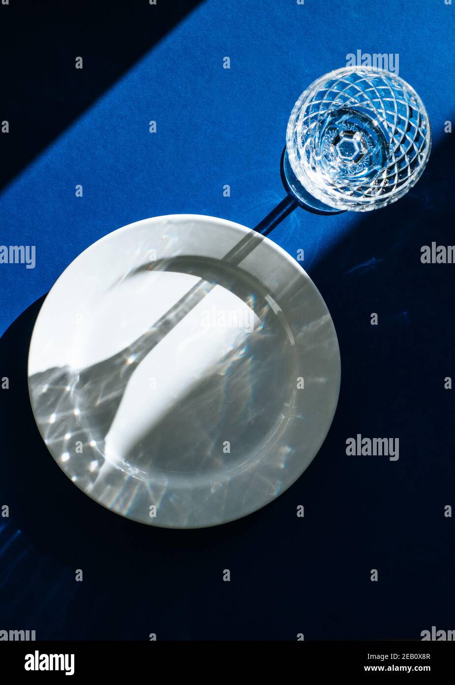 Table avec plaque blanche vide et verre cristal isolé sur fond bleu foncé, ombres sombres et lumière du soleil se reflétant à travers le verre à vin Banque D'Images