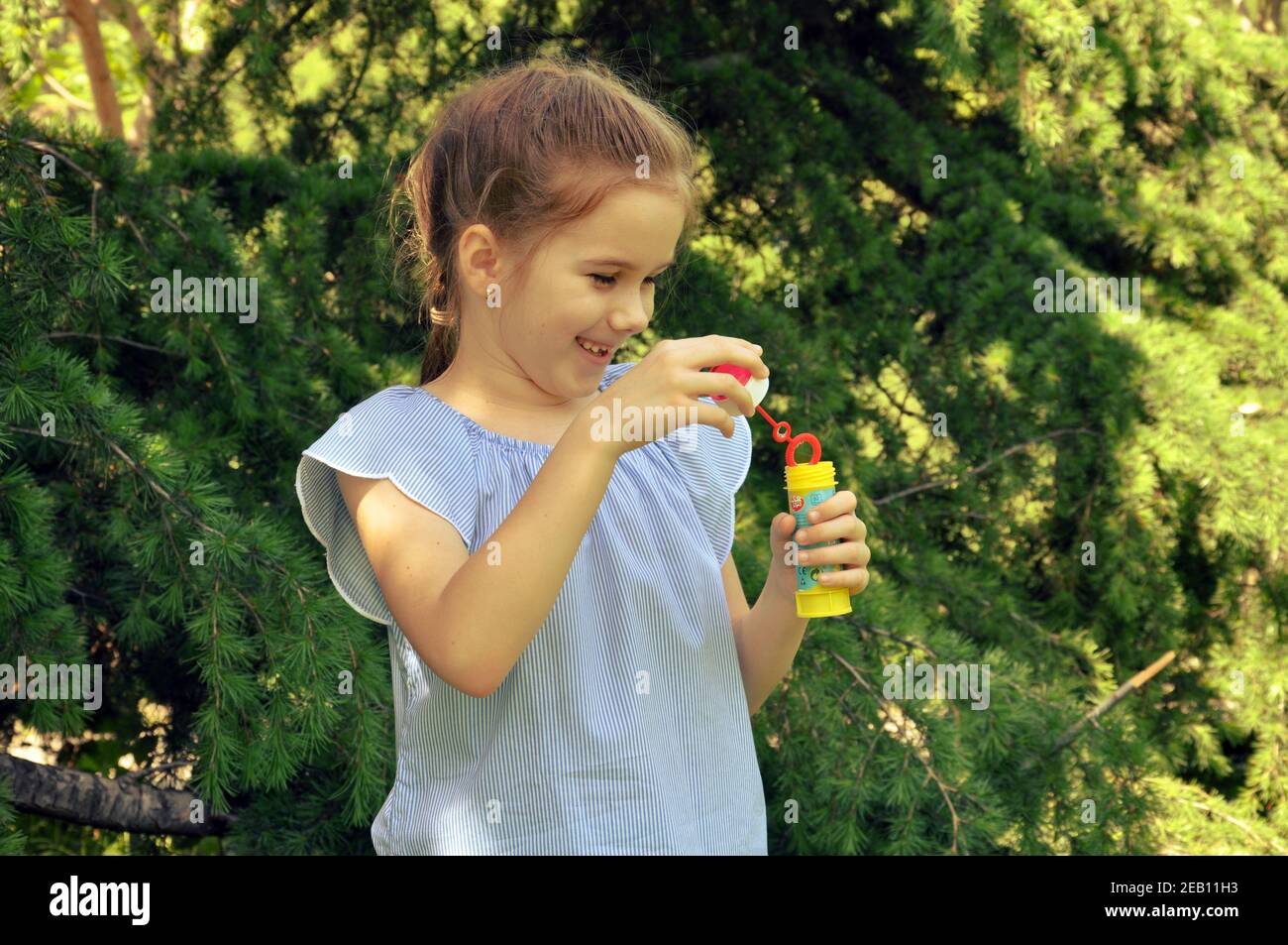 Mignonne fille blonde de cheveux, 7-9, se préparant à souffler les bulles de l'eau dans le parc, vert arbres fond, sélectif foyer, espace de copie Banque D'Images