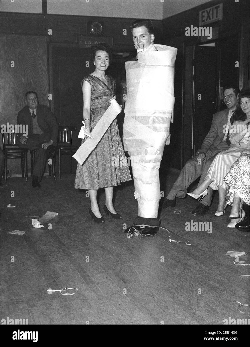 Dans les années 1950, historique, lors d'une fête d'anniversaire dans une salle de réception d'un hôtel, un grand jeune homme debout à côté d'une femme s'est enveloppé de papier marron dans le cou pour se faire un petit cadeau dans sa bouche, un jeu de fête de cette époque sans aucun doute, Angleterre, Royaume-Uni. La dame debout à côté de lui tient un rouleau de papier probablement ayant fait l'acte elle-même. Banque D'Images
