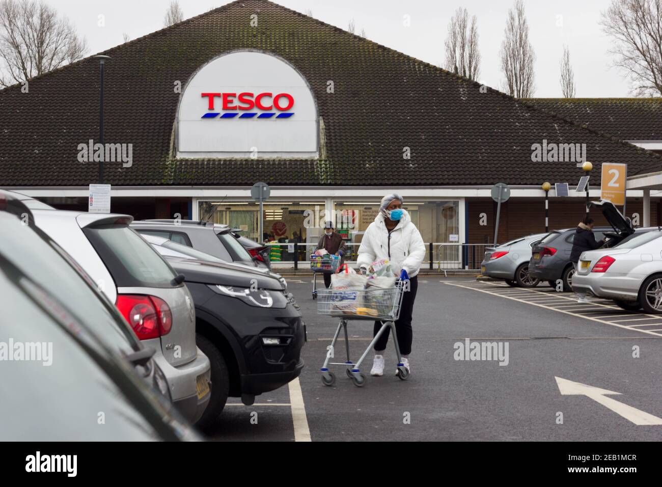 Femme shopper portant un masque de visage poussant chariot à son voiture dans le parking de supermarché Tesco, Kent Banque D'Images