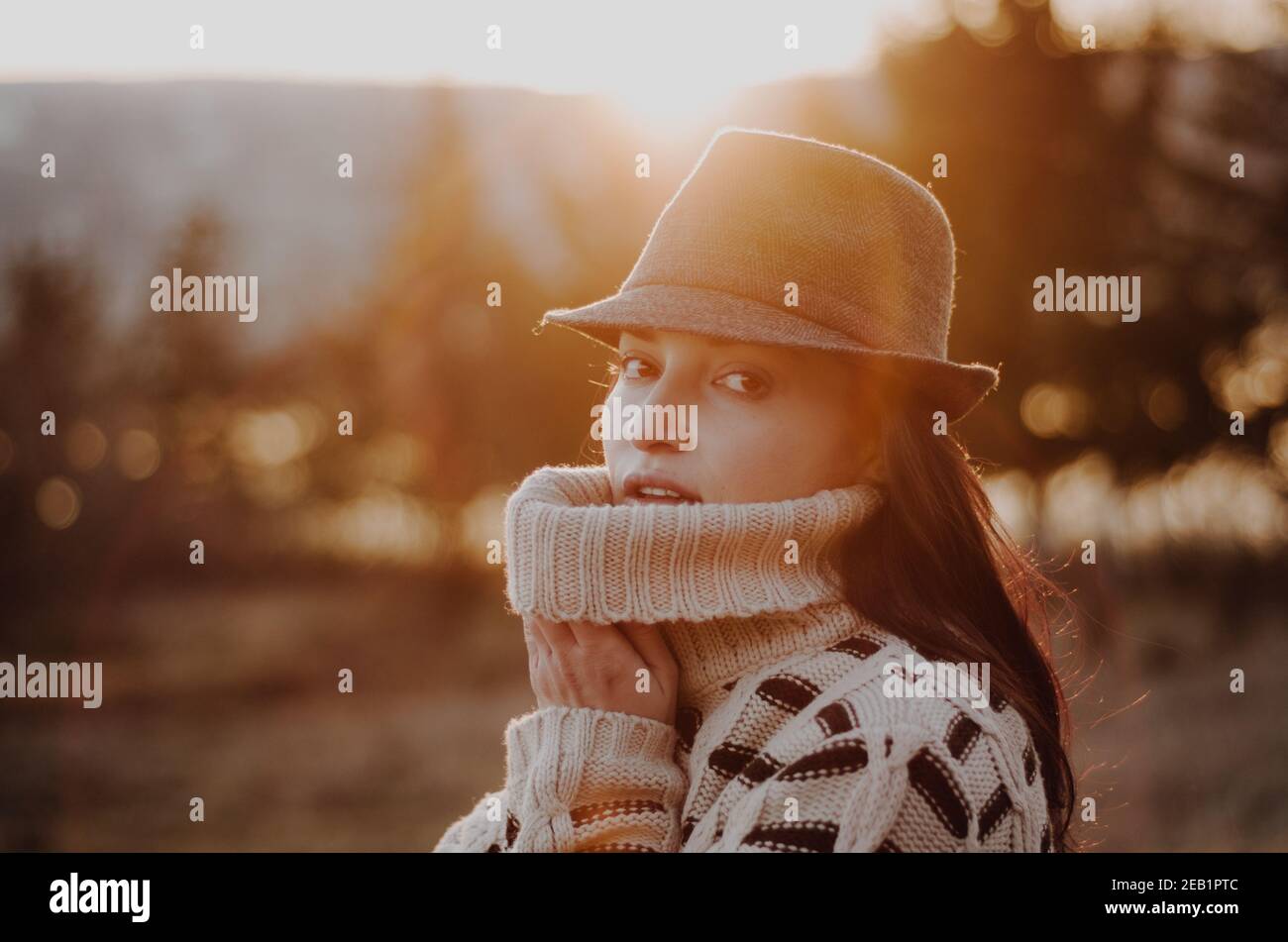 Portrait d'une jeune femme en chandail chaud portant un chapeau dans une atmosphère vibe au coucher du soleil dans les bois avec lac en arrière-plan Banque D'Images