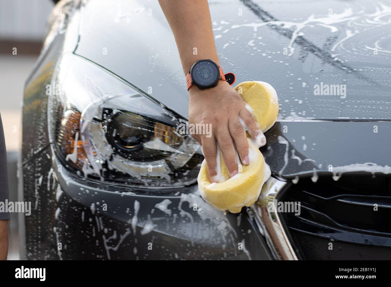 Laver la voiture à la main à l'eau savonneuse. L'homme nettoie la voiture avec une éponge, de l'eau et du détergent. Banque D'Images