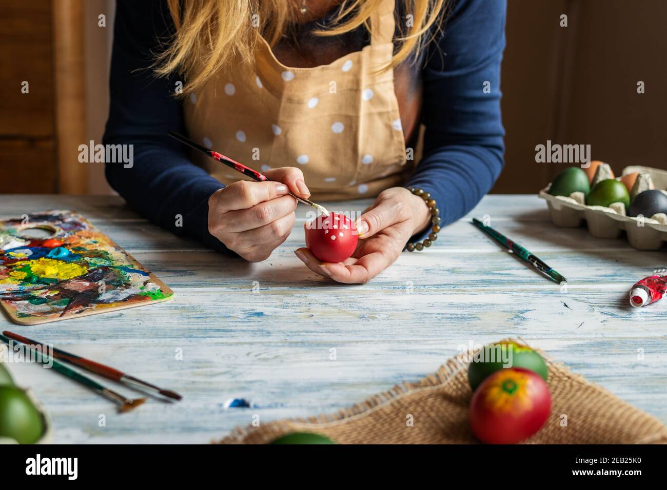 Femme peignant l'œuf et se préparant à la fête de Pâques. Pinceau à main femelle et palette sur table en bois blanc. Décoration à la maison Banque D'Images