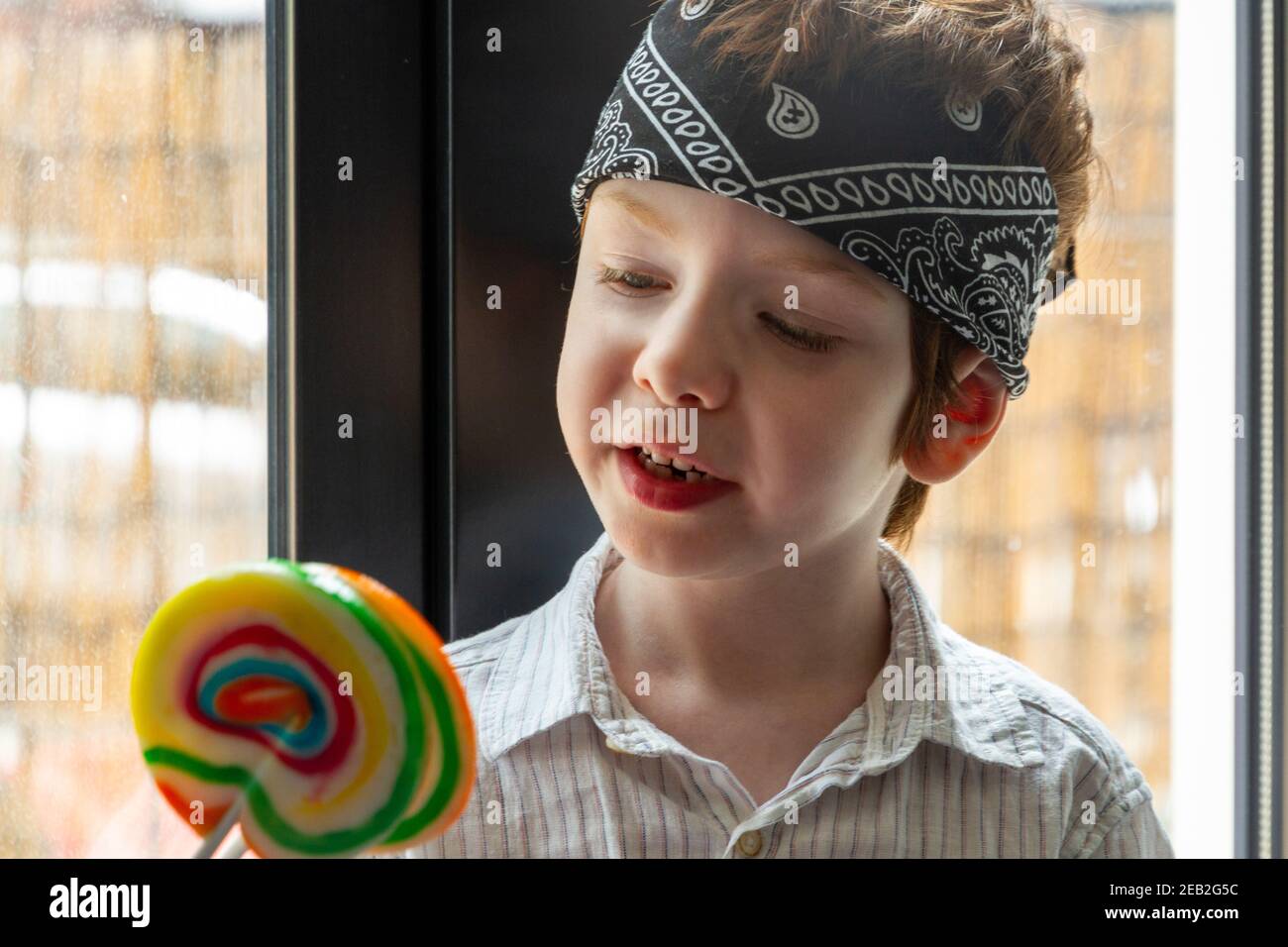 Portrait d'un adorable garçon aux cheveux rouges, aux yeux bleus, regardant un lollipop coloré près d'une fenêtre Banque D'Images
