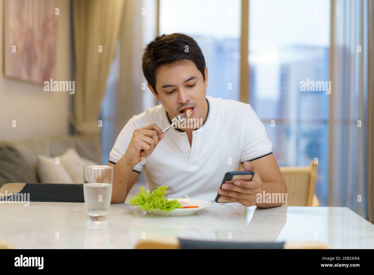 Un homme asiatique prend un petit déjeuner américain tout en étant assis sur une chaise pour utiliser le téléphone, consulter des e-mails ou des informations dans le salon de la maison. Banque D'Images
