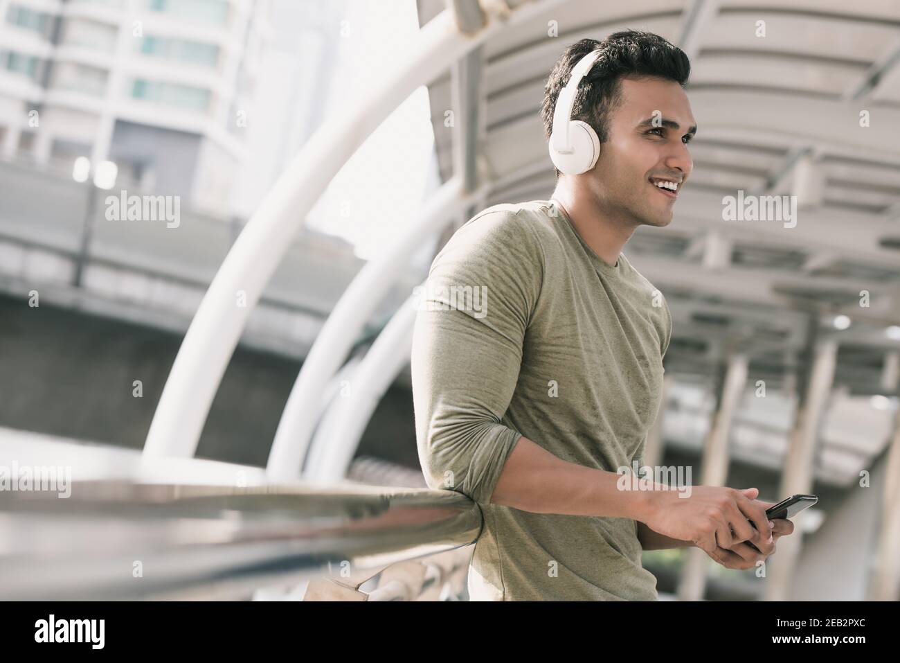Joyeux jeune homme indien élégant portant un casque d'écoute pour écouter de la musique depuis un smartphone en ville Banque D'Images