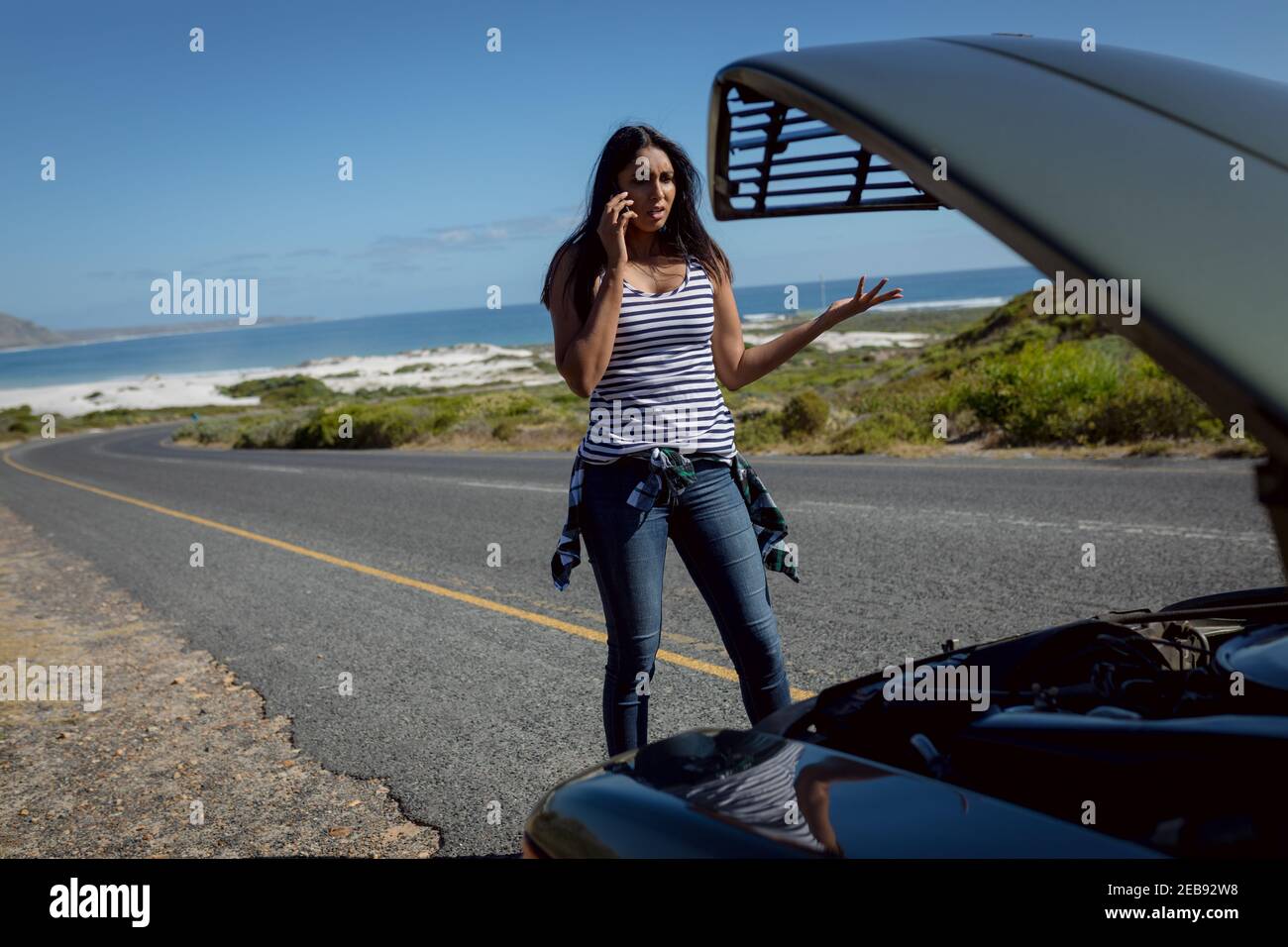 Femme de race mixte parlant avec un smartphone debout sur la route à côté voiture en panne avec capot ouvert Banque D'Images