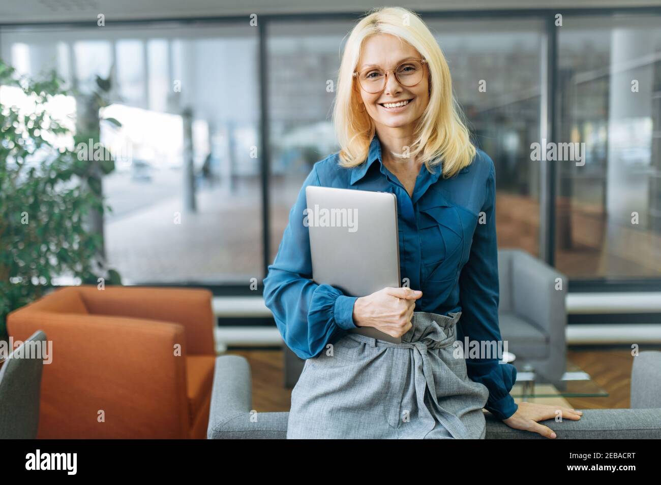 Portrait d'une femme d'affaires heureuse et confiante en eyaglasses et élégante tenue habillée. Femme d'âge moyen qui a réussi à regarder directement la caméra, souriant, tenant l'ordinateur portable dans les bras Banque D'Images