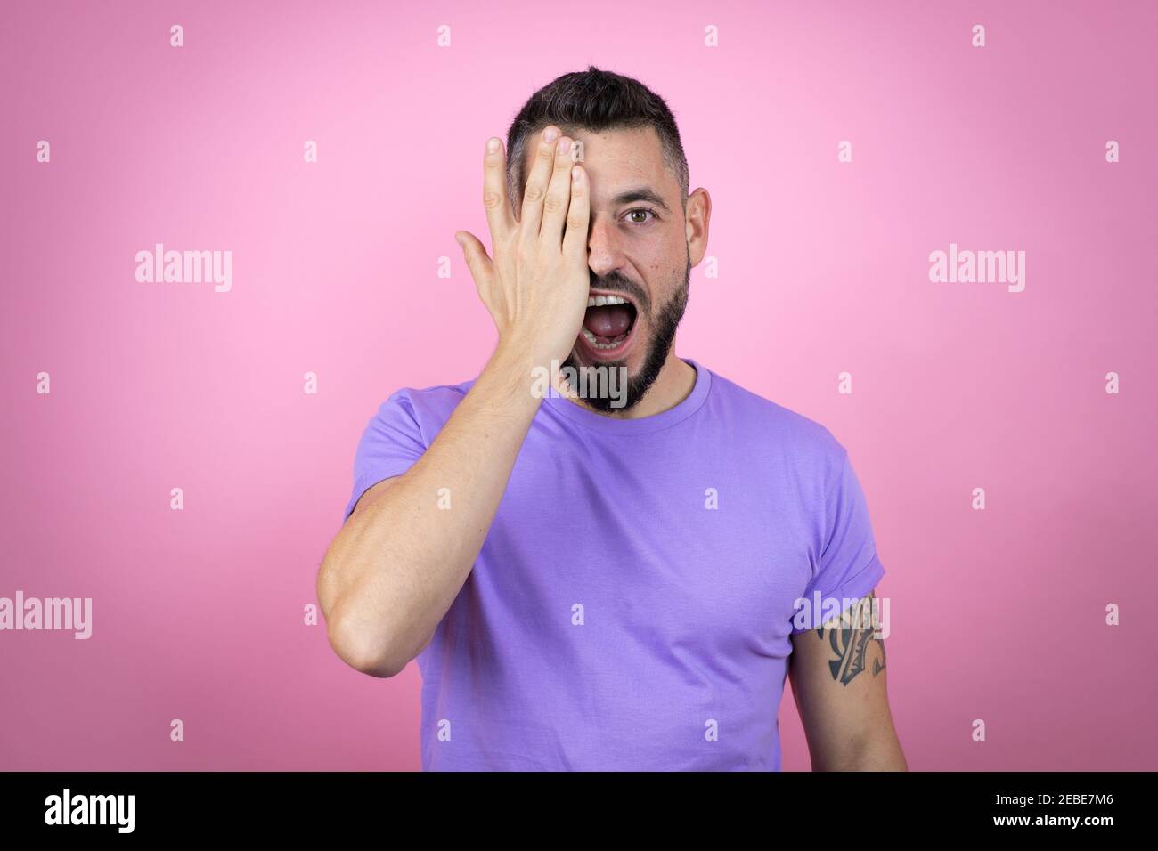 Jeune homme beau portant un t-shirt décontracté sur fond rose couvrant un œil avec la main, un sourire assuré sur le visage et une émotion surprise. Banque D'Images
