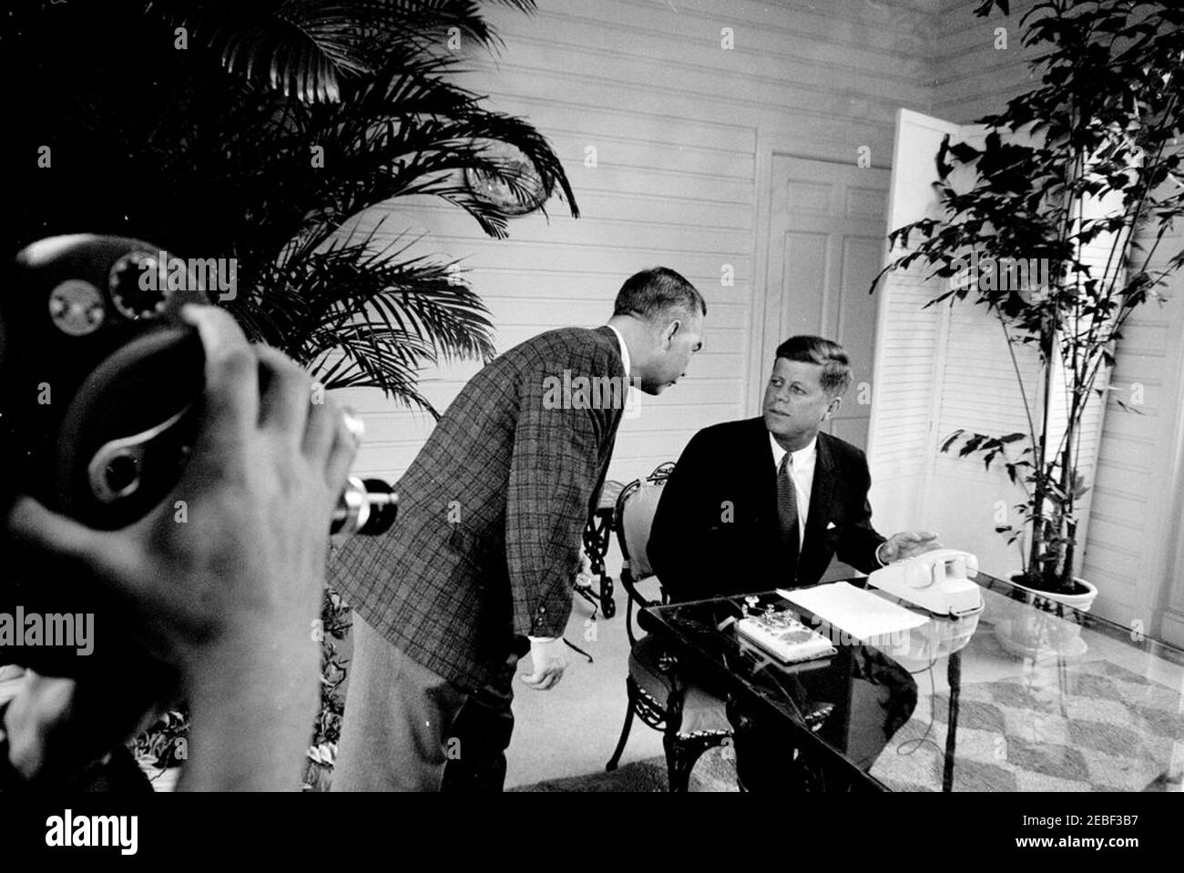 Cérémonie d'ouverture de la foire de Seattle Worldu0027s au départ de Palm Beach. Le président John F. Kennedy (assis à la table du patio) s'entretient avec Robert Bright, de l'American Telephone and Telegraph Company (ATu0026T), avant la cérémonie d'ouverture de la foire internationale de Seattle Worldu0027s en 1962 (également connue sous le nom d'exposition Century 21) depuis la résidence de C. Michael Paul à Palm Beach, en Floride. Un caméraman non identifié (sur le bord le plus éloigné du cadre) filme à gauche. Le président Kennedy a prononcé des remarques par téléphone et a appuyé sur une touche de télégraphe en or (sur la table) pour ouvrir la foire par le signal satellite de Palm Beach. Banque D'Images Cérémonie d'ouverture de la foire de Seattle Worldu0027s au départ de Palm Beach. Le président John F. Kennedy (assis à la table du patio) s'entretient avec Robert Bright, de l'American Telephone and Telegraph Company (ATu0026T), avant la cérémonie d'ouverture de la foire internationale de Seattle Worldu0027s en 1962 (également connue sous le nom d'exposition Century 21) depuis la résidence de C. Michael Paul à Palm Beach, en Floride. Un caméraman non identifié (sur le bord le plus éloigné du cadre) filme à gauche. Le président Kennedy a prononcé des remarques par téléphone et a appuyé sur une touche de télégraphe en or (sur la table) pour ouvrir la foire par le signal satellite de Palm Beach. Banque D'Images