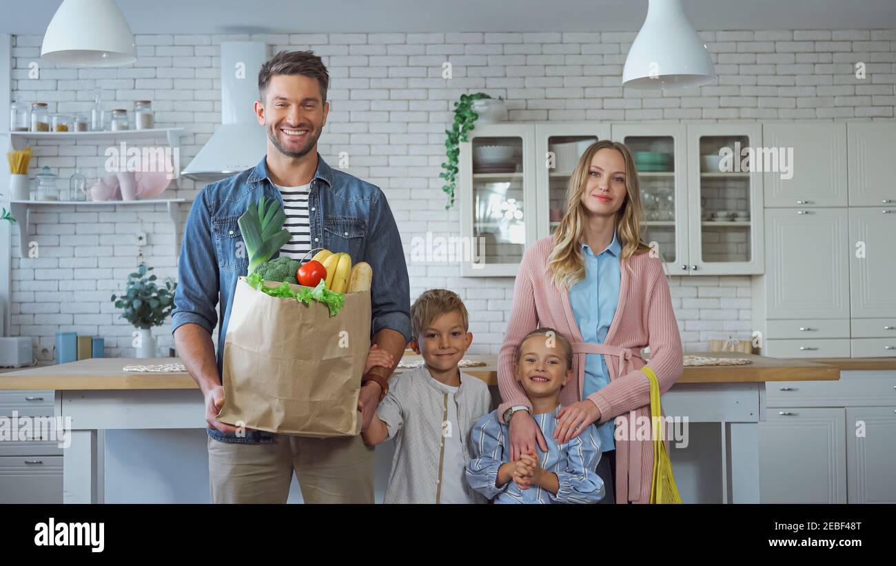 Homme souriant tenant un sac de papier avec des aliments frais près de la famille avec enfants dans la cuisine Banque D'Images