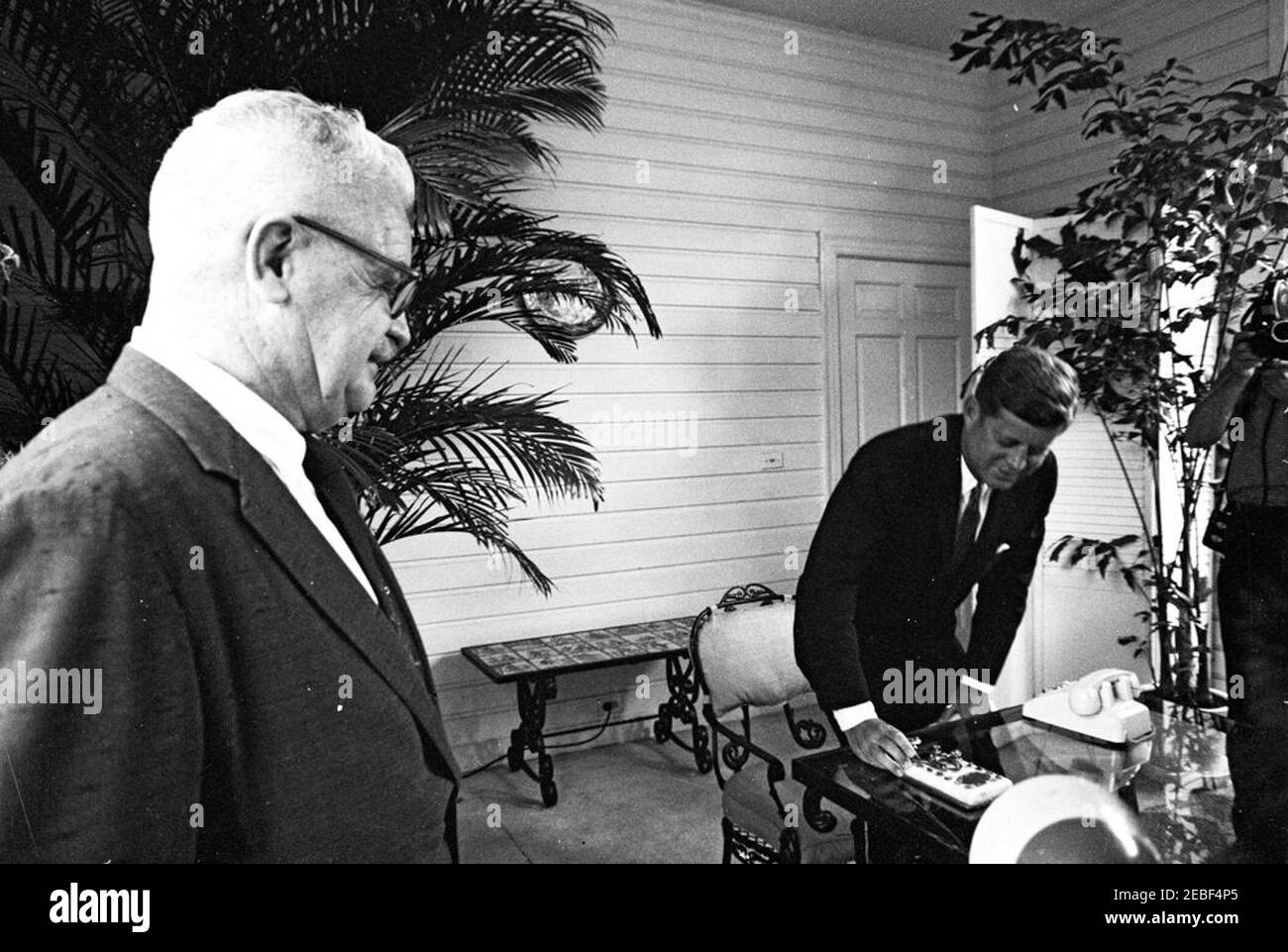 Cérémonie d'ouverture de la foire de Seattle Worldu0027s au départ de Palm Beach. Le président John F. Kennedy (accroupi à la table du patio) appuie sur une touche de télégraphe en or lors de la cérémonie d'ouverture de la foire de Seattle Worldu0027s en 1962 (également connue sous le nom d'exposition Century 21) depuis la résidence de C. Michael Paul à Palm Beach, en Floride. Le chef de l'Agence des communications de la Maison Blanche (WHCA), le colonel George J. McNally, se trouve à gauche; un caméraman non identifié (à l'extrémité du cadre) filme à droite. Le Président Kennedy a prononcé des remarques par téléphone et a appuyé sur la touche du télégraphe pour ouvrir la foire par le biais du signal satellite Banque D'Images Cérémonie d'ouverture de la foire de Seattle Worldu0027s au départ de Palm Beach. Le président John F. Kennedy (accroupi à la table du patio) appuie sur une touche de télégraphe en or lors de la cérémonie d'ouverture de la foire de Seattle Worldu0027s en 1962 (également connue sous le nom d'exposition Century 21) depuis la résidence de C. Michael Paul à Palm Beach, en Floride. Le chef de l'Agence des communications de la Maison Blanche (WHCA), le colonel George J. McNally, se trouve à gauche; un caméraman non identifié (à l'extrémité du cadre) filme à droite. Le Président Kennedy a prononcé des remarques par téléphone et a appuyé sur la touche du télégraphe pour ouvrir la foire par le biais du signal satellite Banque D'Images