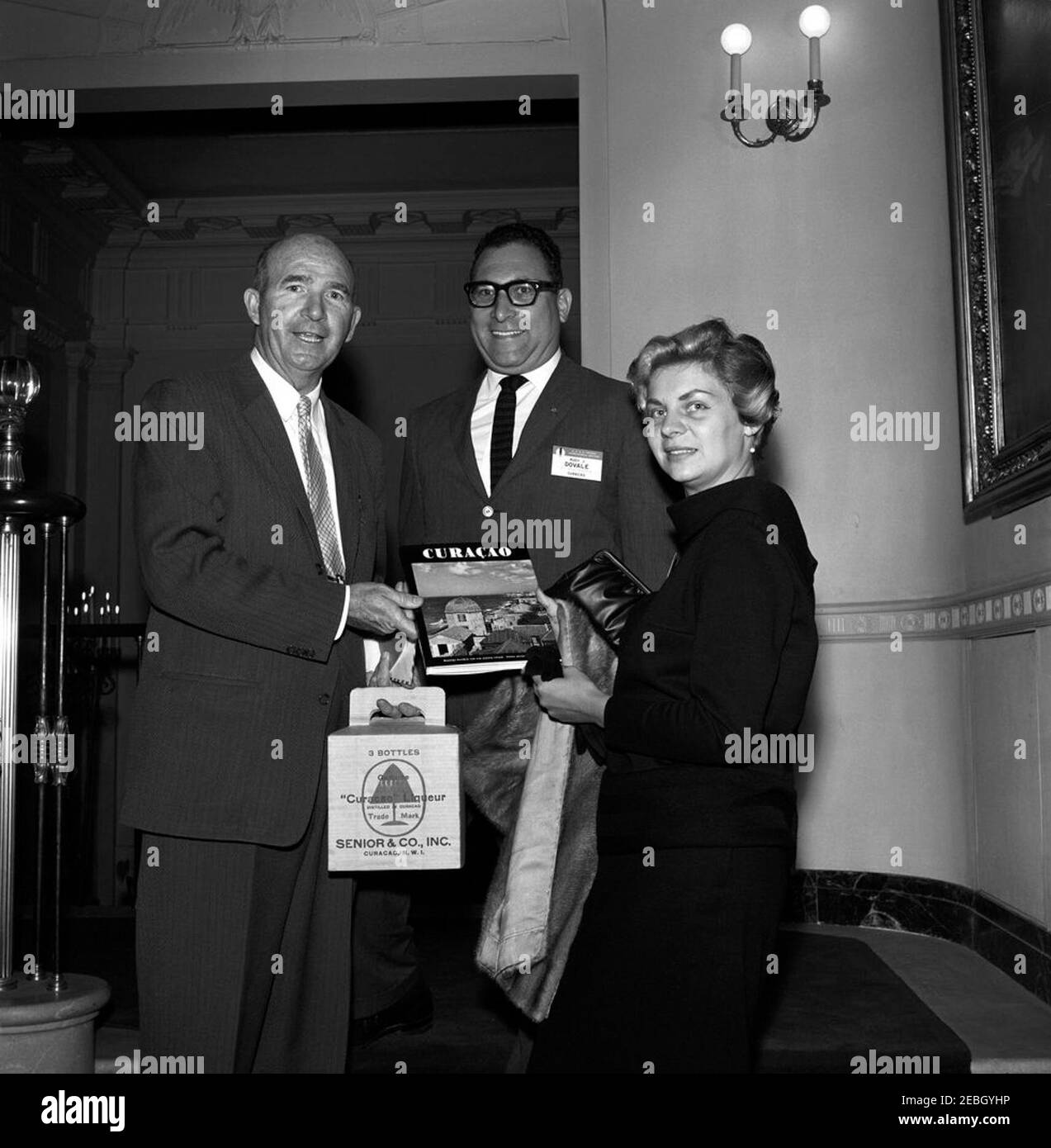 Présentation du livre et de la liqueur de Curau00e7ao pour le président de Rudy J. Dovale (R. J. Dovale Advertising, Curau00e7ao) [Dave Powers]. Rudy J. Dovale (au centre), de R. J. Dovale Advertising à Curau00e7ao, présente à l'adjoint présidentiel Dave Powers (à gauche) un livre et une liqueur de Curau00e7ao pour le président John F. Kennedy. La femme de droite n'est pas identifiée. Grand escalier, hall d'entrée, Maison Blanche, Washington, D.C. Banque D'Images