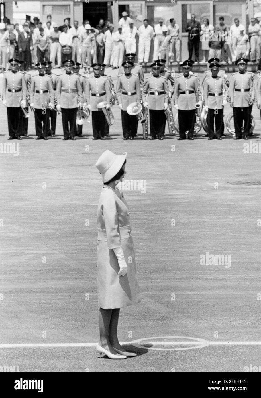 Voyage au Mexique : cérémonies d'arrivée, aéroport international de Mexico, Mexico, Mexique, 11:00. La première dame Jacqueline Kennedy assiste à une cérémonie d'arrivée en l'honneur du président John F. Kennedy et de Mme Kennedy à Mexico. Les membres d'un groupe se tiennent en arrière-plan. Aéroport international Benito Juu00e1rez, Mexico, Mexique. Banque D'Images