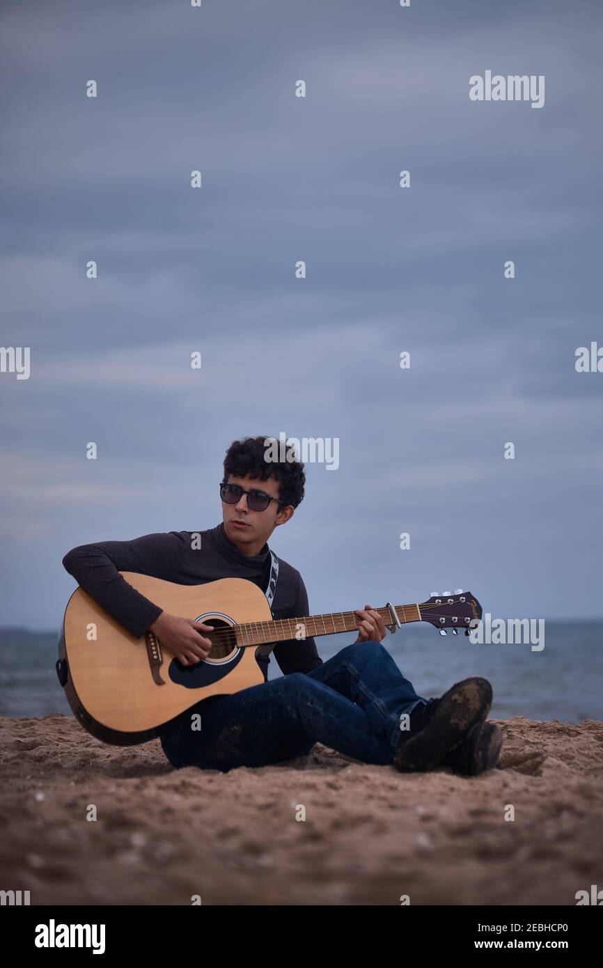 jeune adolescent caucasien jouant de la guitare acoustique sur la plage. lunettes de soleil et vêtements foncés. Banque D'Images