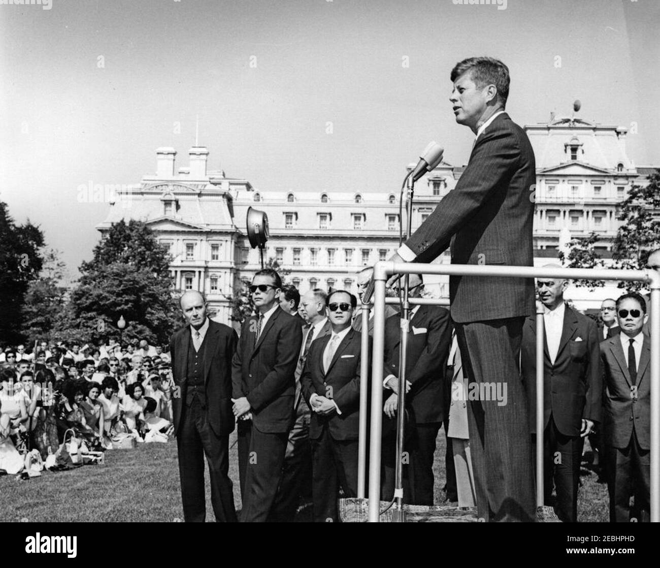 Visite des étudiants du service après-vente américain, 10:00. Le président John F. Kennedy (à droite, à micros) fait des remarques de la plate-forme Speakersu0027 à un groupe d'étudiants de l'American Field Service (AFS) réunis sur la pelouse sud de la Maison Blanche, Washington, D.C. debout à l'arrière, à gauche de la plate-forme (de gauche à droite): Non identifié; Agent du service secret de la Maison Blanche, Bill Payne; ministre de l'ambassade du Pakistan, Mohammed Masood (à l'arrière, partiellement caché); non identifié; ambassadeur de Malaya, Omar Ong Yoke Lin (portant des lunettes de soleil); non identifié; ambassadeur d'Australie, Sir Howard be Banque D'Images