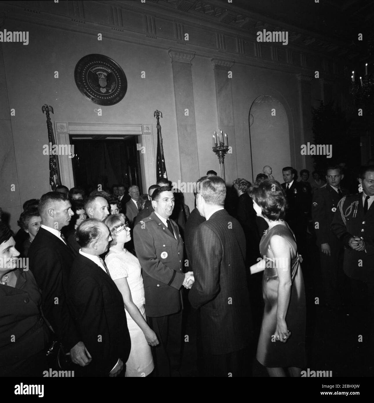 Fête de Noël du personnel de la Maison Blanche dans la salle est, 6:30. Le président John F. Kennedy et la première dame Jacqueline Kennedy saluent les invités rassemblés à Cross Hall, lors d'une fête de Noël pour le personnel de la Maison Blanche; le président Kennedy (au centre à droite, de retour à la caméra) serre la main avec le dirigeant de l'Agence de communication de la Maison Blanche (WHCA), le major Anthony S. Suglia; Mme Kennedy (en profil) tremble la main avec un autre invité caché derrière le Président. L'aide militaire au Président général Chester V. Clifton se trouve à l'extrême droite ; les clients remplissent le couloir et entrent dans la salle bleue à gauche. White House, Washington Banque D'Images