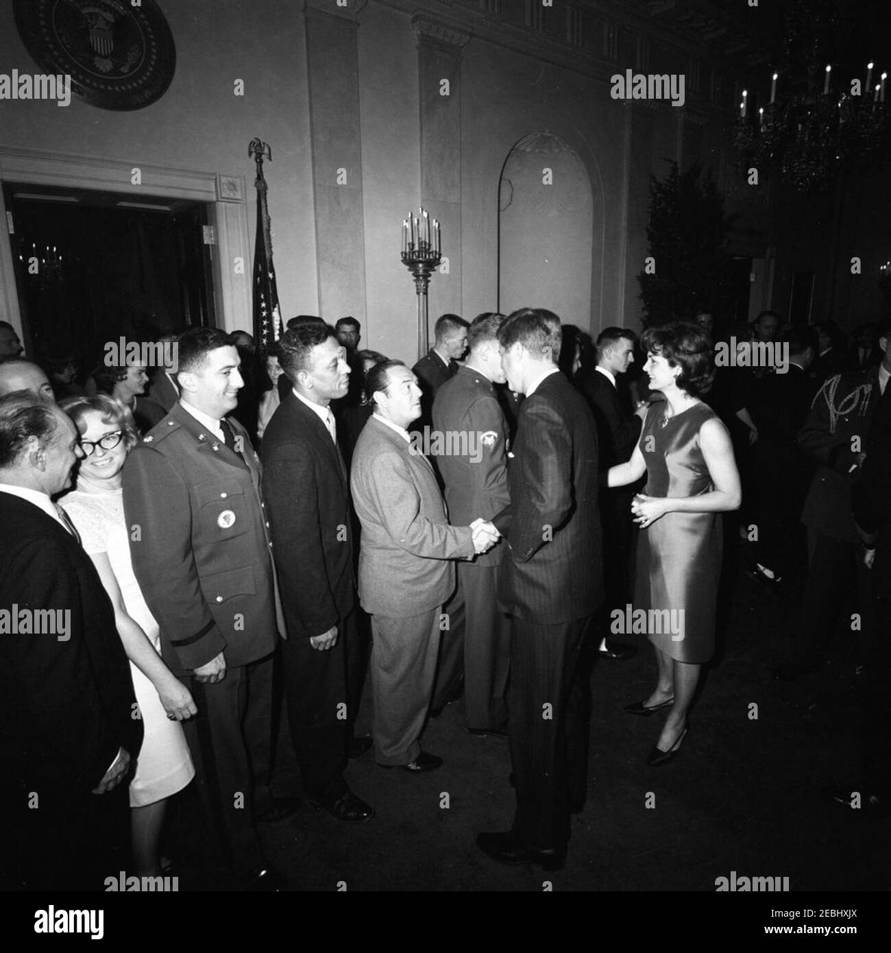 Fête de Noël du personnel de la Maison Blanche dans la salle est, 6:30. Le président John F. Kennedy et la première dame Jacqueline Kennedy saluent les invités rassemblés à Cross Hall, lors d'une fête de Noël pour le personnel de la Maison Blanche ; le président Kennedy (au centre à droite, de retour à la caméra) serre la main avec l'électricien du réseau de médias de la Maison Blanche, Cleve Ryan ; Mme Kennedy (en profil) tremble la main avec un autre invité qui est partiellement caché derrière le Président. Le major Anthony S. Suglia, officier de l'Agence des communications de la Maison Blanche (WHCA), se tient deux personnes derrière M. Ryan; les clients remplissent le couloir et entrent dans la salle bleue à gauche. Blanc Banque D'Images