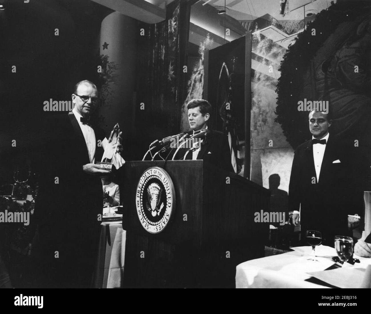 Banquet de remise des prix de la Fondation Kennedy, 8:49PM. Le président John F. Kennedy (sous la lecterne) présente les prix lors du dîner de la Joseph P. Kennedy, Jr. Foundation First International Awards pour ses réalisations en recherche, en service et en leadership dans le domaine du retard mental. John G. Fettinger, récipiendaire du prix et président de l'Association nationale pour les enfants retardés, se trouve à gauche; Sargent Shriver, directeur du corps de la paix et directeur exécutif de la Fondation, se trouve à droite. Statler Hilton Hotel, Washington, D.C. Banque D'Images