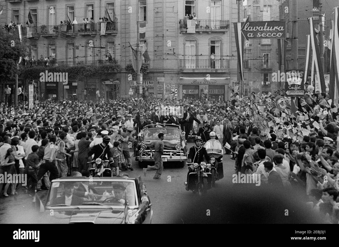 Voyage en Europe: Italie, Naples: Cortège, 5:40. Le président John F. Kennedy (debout dans une deuxième voiture) déonde d'un cabriolet ouvert tandis que son cortège voyage à travers Naples, Italie. Le président de l'Italie, Antonio Segni, se présente avec le président Kennedy. Également en photo : assistant spécial du président Kennedy, Kenneth P. Ou2019Donnell; agents du Service secret de la Maison Blanche, Dave Grant, James J. Rowley et Roy Kellerman. Un accompagnateur de moto voyage avec le cortège de voiture; la foule suit les rues. Banque D'Images