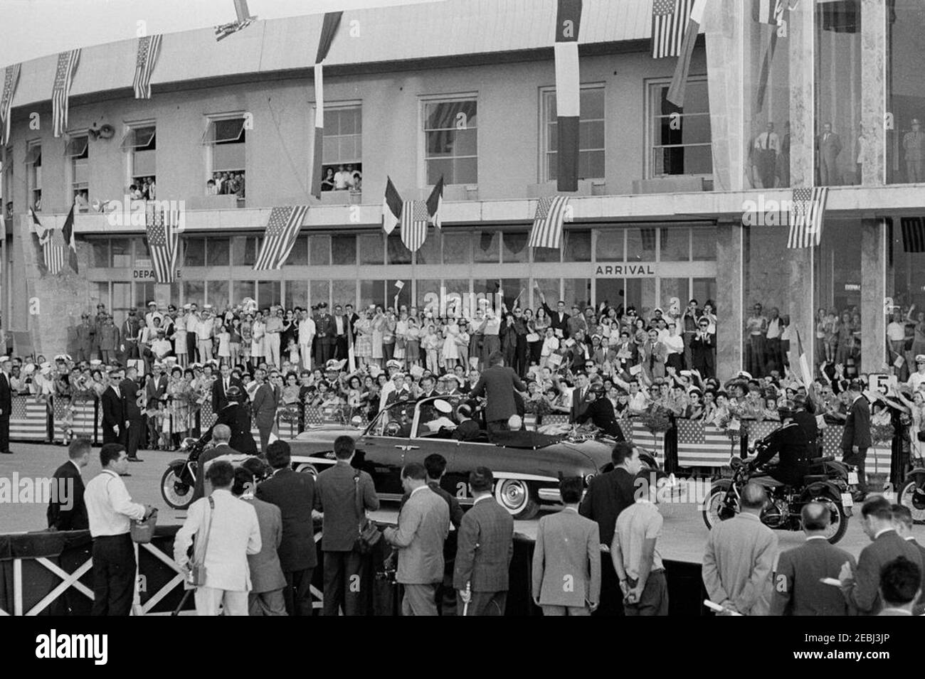 Voyage en Europe : Italie, Naples : départ du Président Kennedyu2019s, 6:30. Le président John F. Kennedy (debout en voiture, face à la caméra) se déporte devant une foule d'un cabriolet ouvert à son arrivée à l'aéroport Capodichino de Naples, en Italie ; le président Kennedy a prononcé des remarques à l'aéroport avant de partir pour les États-Unis. Le président de l'Italie, Antonio Segni, est assis dans la voiture à côté du président Kennedy. Également en photo : agents du service secret de la Maison Blanche, Win Lawson et Emory Roberts. Banque D'Images