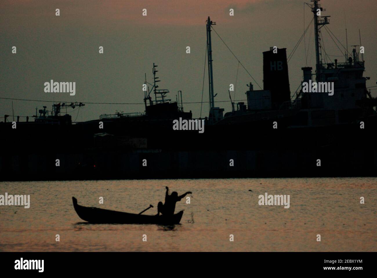 Silhouettes d'une partie d'un navire amarré et d'un pêcheur travaillant avec des filets de poisson d'un bateau flottant sur une piscine intérieure, avant le coucher du soleil dans la zone côtière de Marunda, Jakarta, Indonésie. Banque D'Images