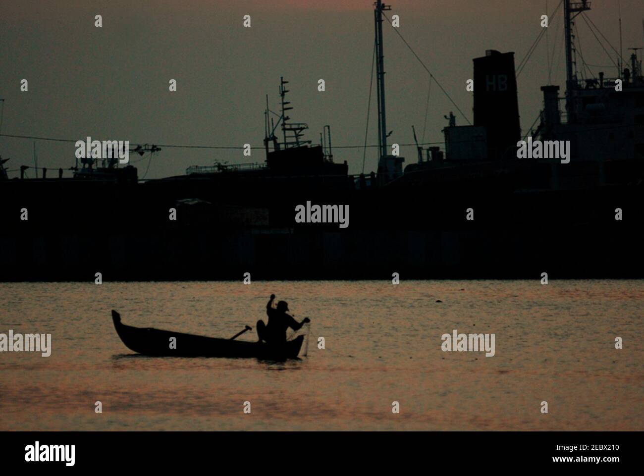 Une partie d'un navire amarré est silhoueted contre la lumière de l'après-midi, en avant-plan d'un pêcheur travaillant avec des filets de poisson d'un bateau flottant sur une piscine intérieure dans la zone côtière de Marunda, Jakarta, Indonésie. Banque D'Images
