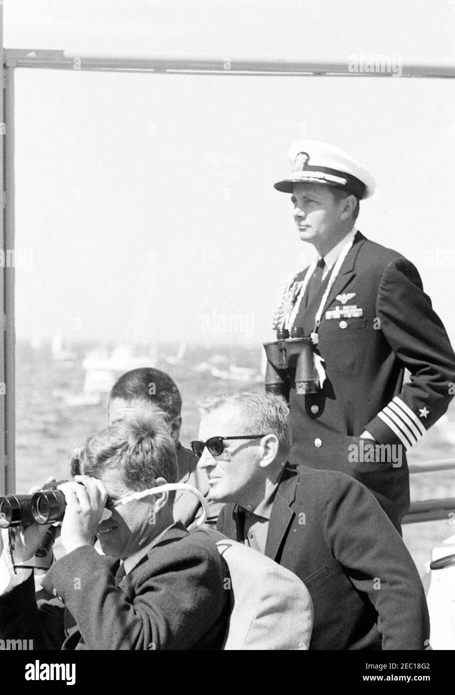 Le Président Kennedy Watches la 1ère course de la coupe Americau0027s. Le président John F. Kennedy (en regardant à travers des jumelles) regarde la première course de la coupe Americau0027s 1962 à bord de l'USS Joseph P. Kennedy, Jr. Également en photo : le sénateur Ben Smith du Massachusetts (pour la plupart caché derrière le président Kennedy); le sous-secrétaire de la Marine, Paul u0022Redu0022 Fay (assis au centre); Aide navale au président, le capitaine Tazewell Shepard (debout). Newport, Rhode Island. Banque D'Images