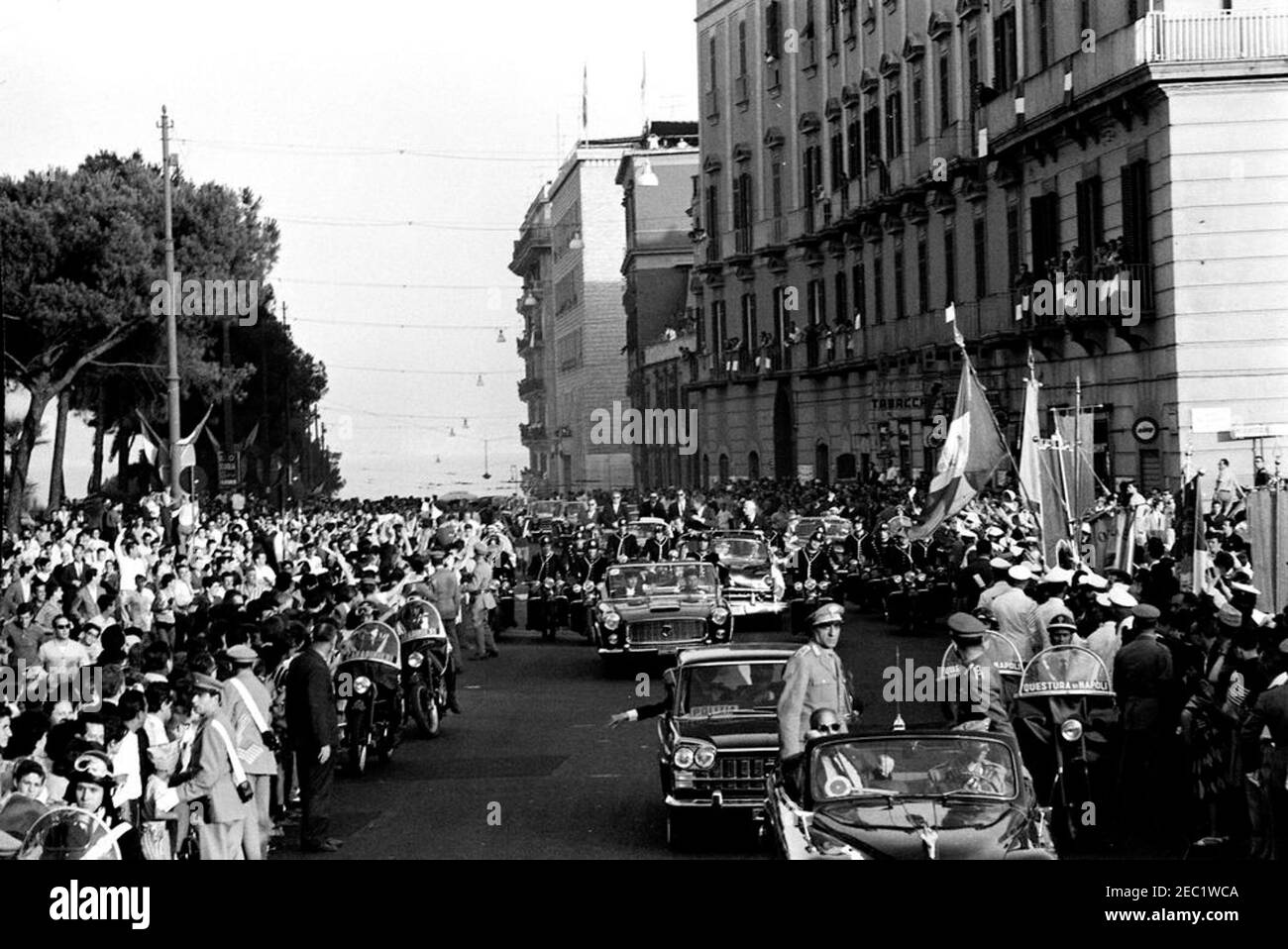 Voyage en Europe: Italie, Naples: Cortège, 5:40. Le président John F. Kennedy (debout en quatrième voiture) fait des vagues d'un cabriolet ouvert alors que son cortège voyage à travers Naples, en Italie. Le président de l'Italie, Antonio Segni, se présente avec le président Kennedy. Également en photo : assistant spécial du président Kennedy, Kenneth P. Ou2019Donnell; agents du service secret de la Maison Blanche, Floyd Boring, James J. Rowley et Roy Kellerman. Un accompagnateur de moto voyage avec le cortège de voiture; la foule suit les rues. Banque D'Images