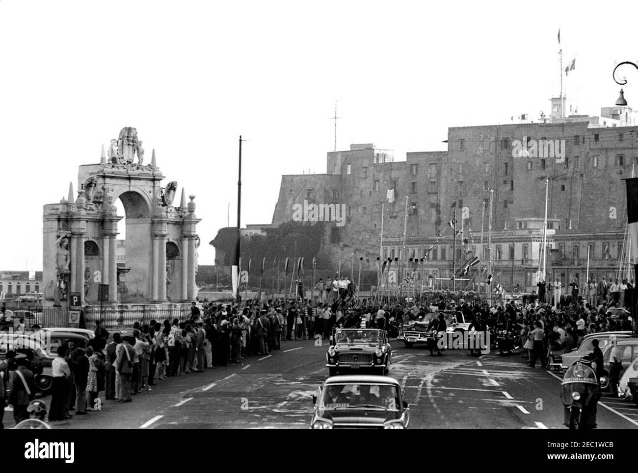 Voyage en Europe: Italie, Naples: Cortège, 5:40. Le président John F. Kennedy (debout dans une troisième voiture) fait des vagues d'un cabriolet ouvert alors que son cortège voyage à travers Naples, Italie. Le président de l'Italie, Antonio Segni, se présente avec le président Kennedy. La Fontana del Gigante, ou Fontaine du géant, se trouve sur la gauche. Un accompagnateur de moto voyage avec le cortège de voiture; la foule suit les rues. Banque D'Images