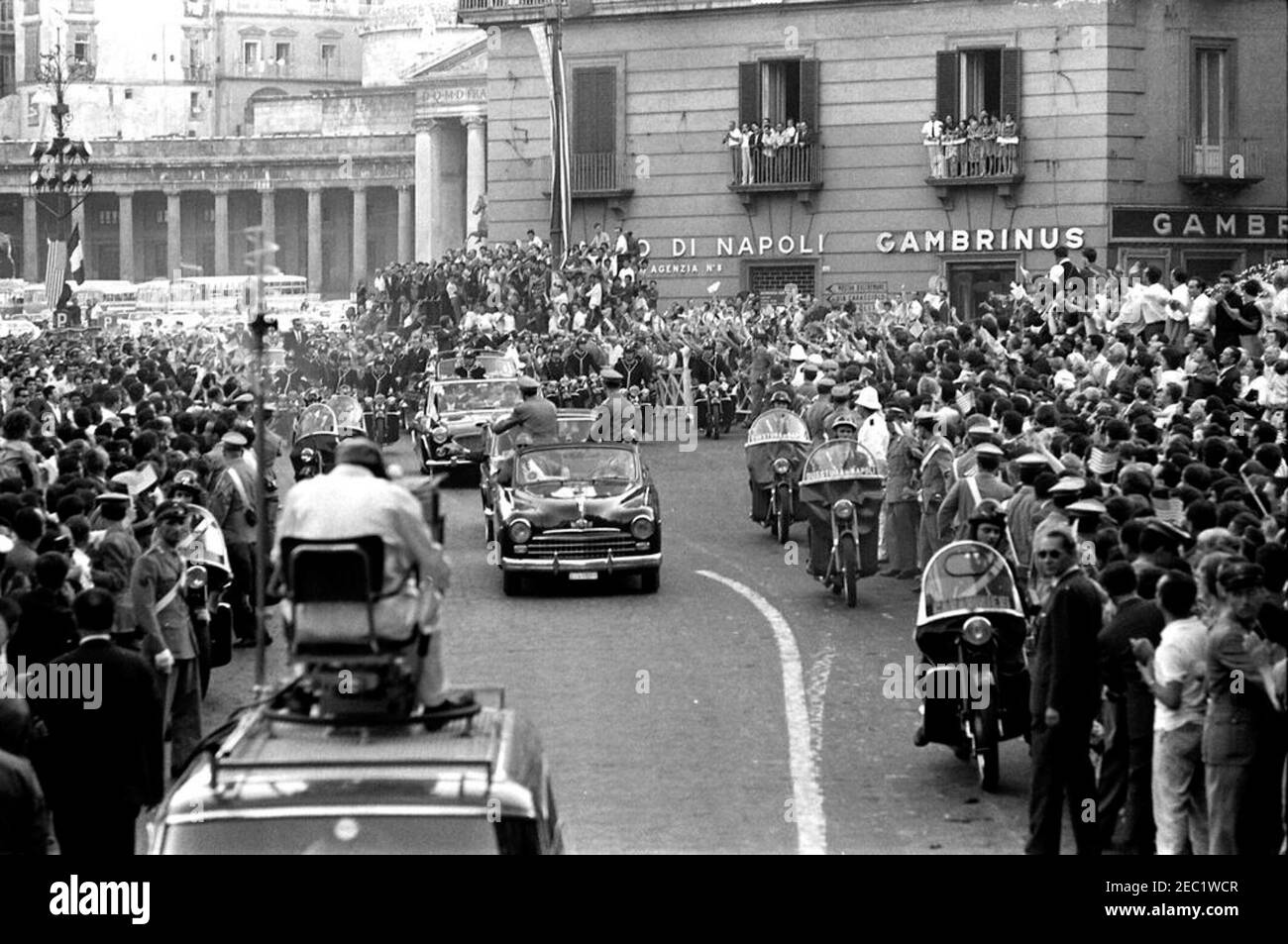 Voyage en Europe: Italie, Naples: Cortège, 5:40. Le président John F. Kennedy (en cinquième voiture) fait des vagues d'un cabriolet ouvert alors que son cortège voyage à travers Naples, en Italie. Le président de l'Italie, Antonio Segni, se présente avec le président Kennedy. La Piazza del Plebiscito est visible en arrière-plan. Un accompagnateur de moto voyage avec le cortège de voiture; la foule suit les rues. Banque D'Images