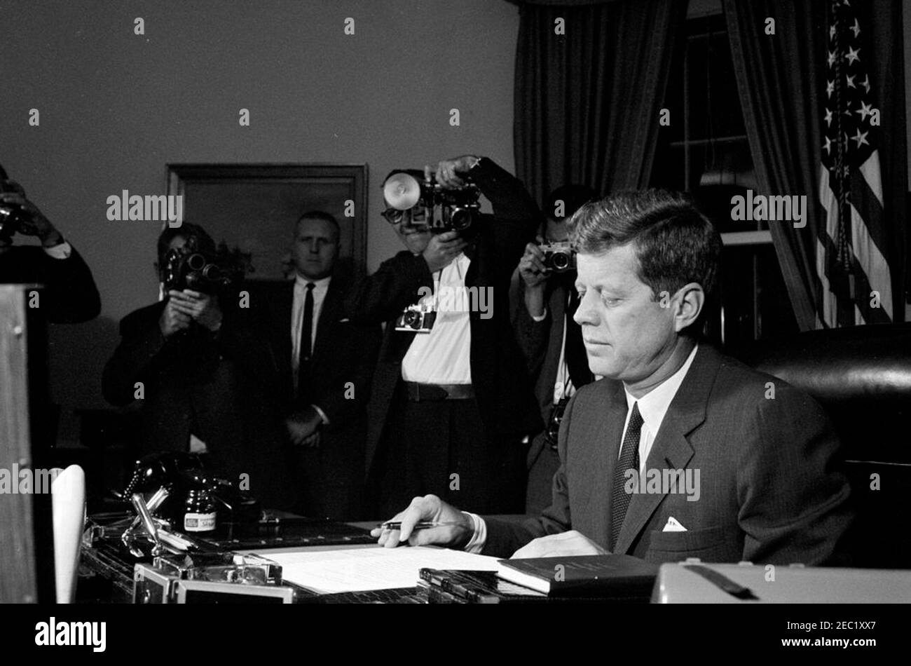Signature de la proclamation, interdiction de la livraison d'armes offensives à Cuba, 7:05. Le Président John F. Kennedy signe l'interdiction de la livraison d'armes offensives à Cuba. Frank Yeager, agent du service secret de la Maison Blanche, se tient contre le mur à gauche ; les photographes ne sont pas identifiés. Oval Office, Maison Blanche, Washington, D.C. Banque D'Images