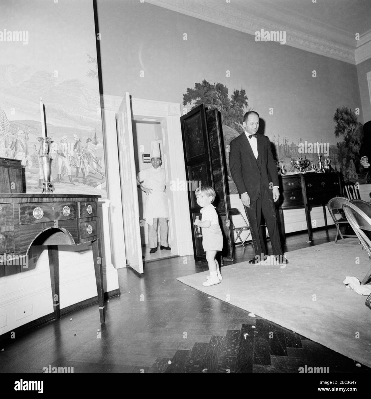 Fête d'anniversaire pour Caroline Kennedy et John F. Kennedy, Jr. John F. Kennedy, Jr. (Tenant une bouteille de Coca-Cola), se tient dans la salle à manger du Presidentu2019s (résidence) de la Maison Blanche, Washington, D.C., lors d'une fête d'anniversaire conjointe pour lui et sa sœur, Caroline Kennedy. Également photographié : assistant chef de la Maison Blanche, Julius Spessot (debout à la porte) ; maître d'hôtel de la Maison Blanche, Eugene Allen. [Les rayures et les imperfections de l'image sont d'origine sur le négatif.] Banque D'Images