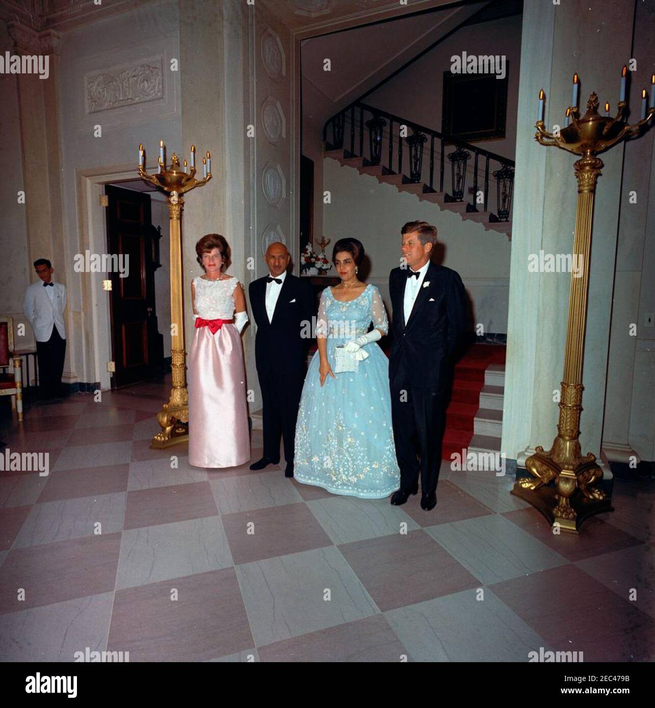 Dîner en l'honneur du roi Mohammad Zahir Shah d'Afghanistan, 8:00. Le Président John F. Kennedy assiste à un dîner en l'honneur du Roi Mohammad u1e92u0101hir Shu0101h et de la Reine Humaira Begum d'Afghanistan. Debout devant le Grand escalier (G-D) : la sœur du président Kennedyu2019s, Eunice Kennedy Shriver; le roi u1e92u0101hir; la reine Humaira; le président Kennedy. Hall d'entrée, Maison Blanche, Washington, D.C. Banque D'Images