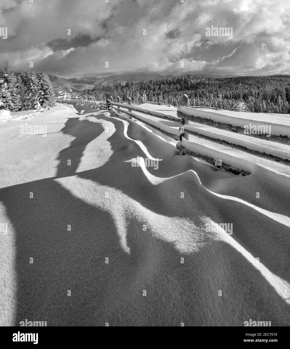 Niveaux de gris. Des ombres ondulées pittoresques sur la neige de la clôture en bois. Montagne alpine hiver hameau en périphérie, sentier enneigé, forêt de sapins. Image haute résolution W Banque D'Images