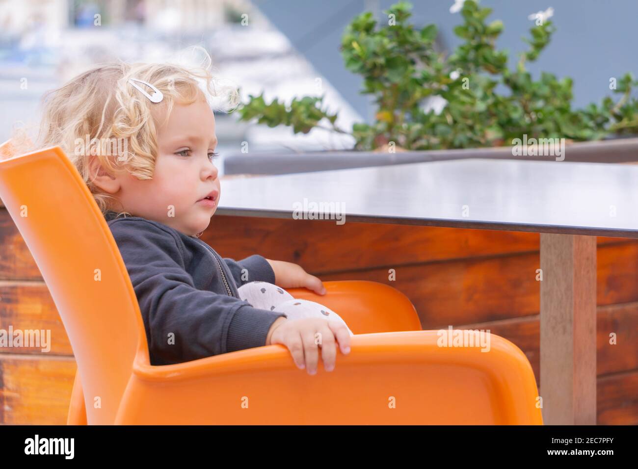 Joyeux petit enfant en bonne santé assis dans un fauteuil orange, adorable petite fille blonde attendant la nourriture dans le café à l'extérieur Banque D'Images